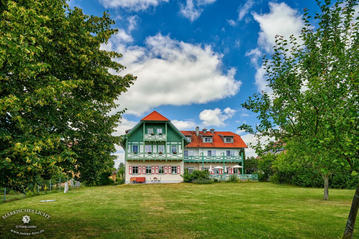 Mitten im Grünen, das Seehotel Huberhof am Oberuckersee in Seehausen. Unten rechts, bei Bedarf beschirmt, die Terrasse zum Frühstücken oder Verweilen und essen. Im Innenhof gibt es dazu einen weiteren Platz.